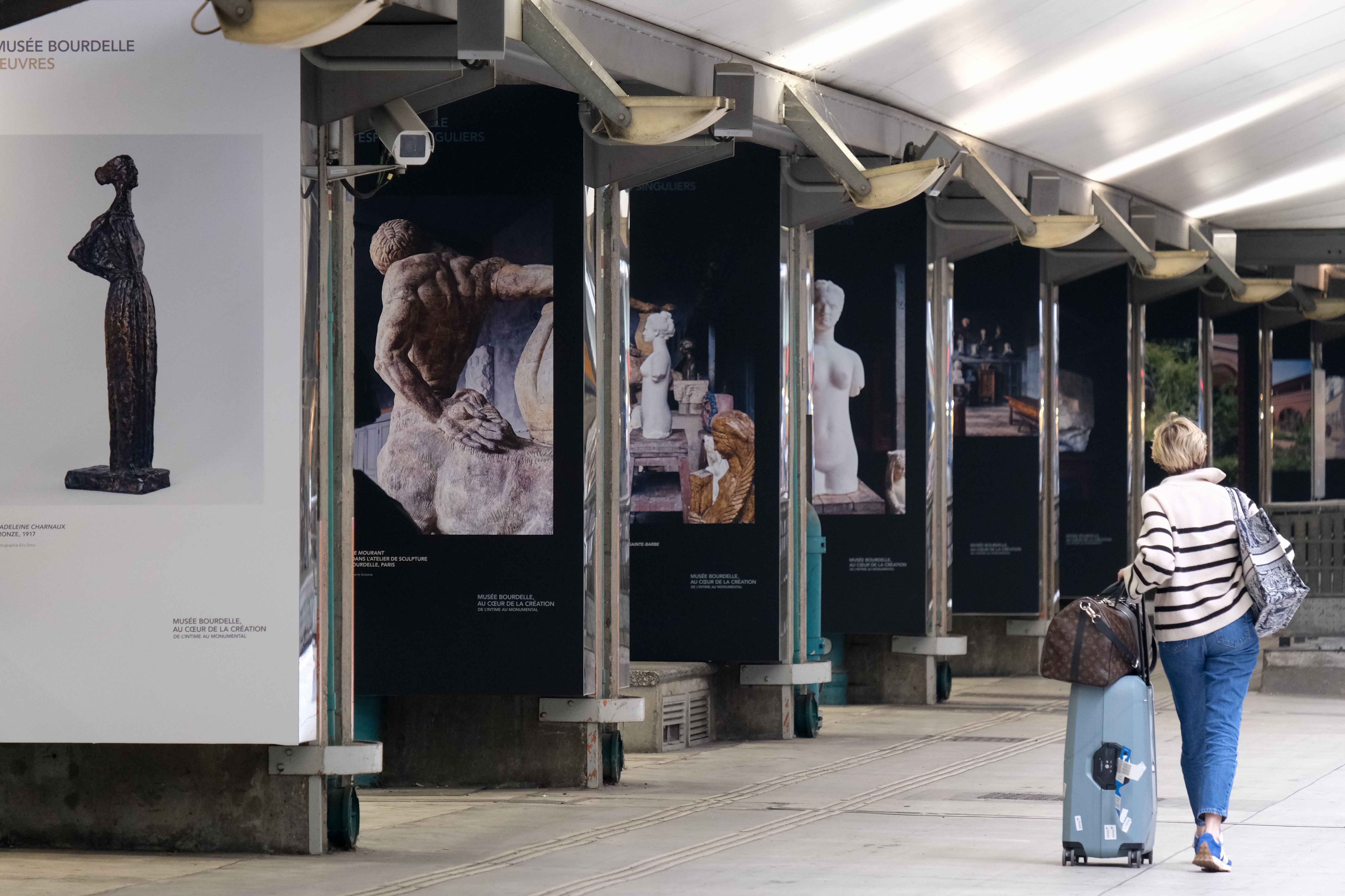 Une rangée de panneaux d'exposition du musée Bourdelle dans la gare de Paris Montparnasse. Une femme avec une valise et 2 sacs longe les panneaux.