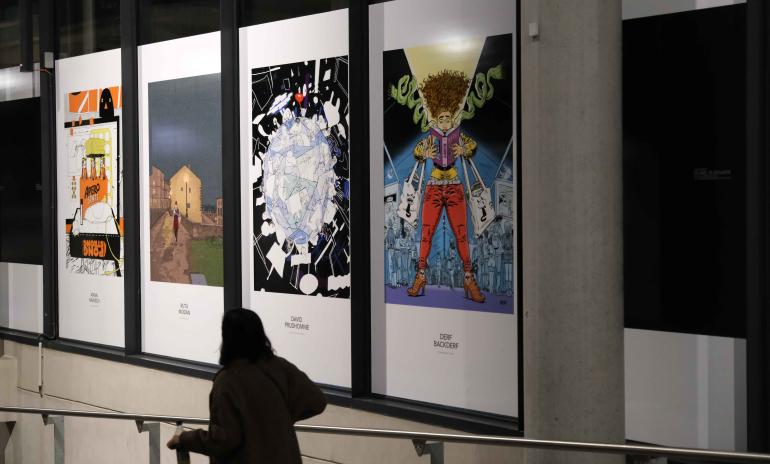 Photo d'une femme avec une valise qui passe devant les panneaux de 4 illustrations de l'expo "50 ans, 50 regards".