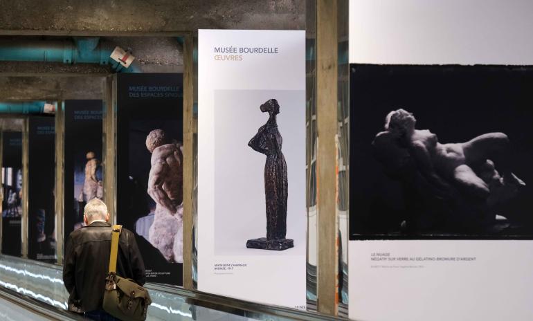 6 panneaux d'expositions qui représentent des sculptures du Musée Bourdelle, positionnés le long d'un tapis roulant sur lequel se trouve un homme avec un sac sur l'épaule.